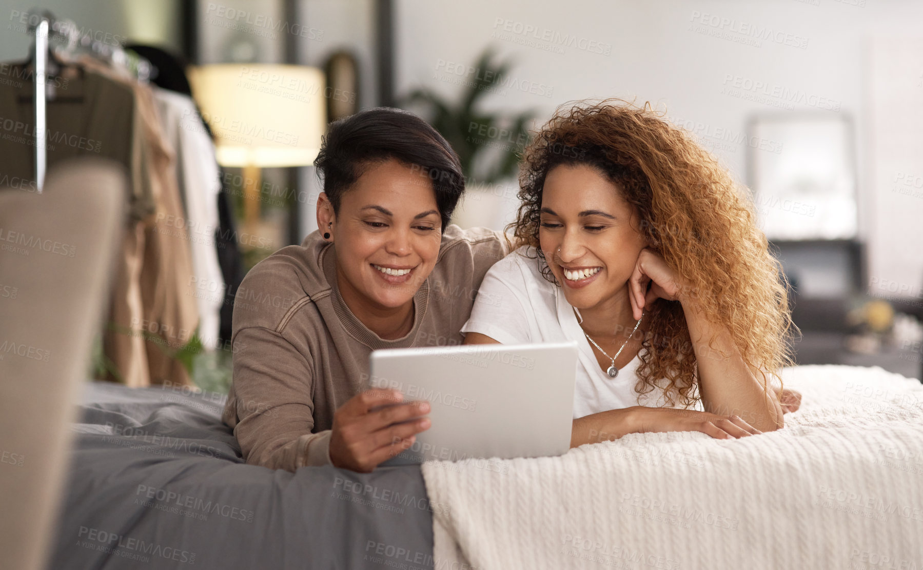 Buy stock photo Shot of a young lesbian couple using a tablet while relaxing in their bedroom