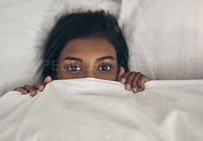 Buy stock photo Portrait of a playful young woman hiding under the covers in a bed