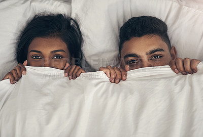 Buy stock photo Portrait of a playful young couple hiding under the covers in a bed