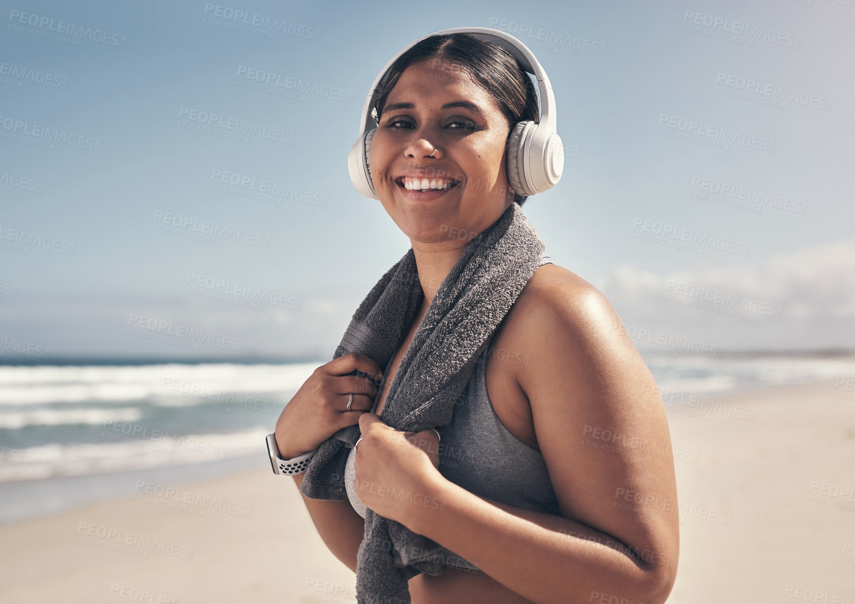 Buy stock photo Shot of a sporty young woman wearing headphones and a towel around her neck on the beach