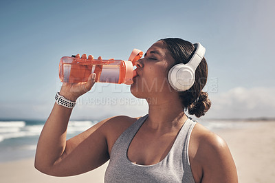 Buy stock photo Shot of a woman wearing headphones and drinking water while out for a workout on the beach