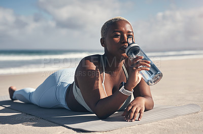 Buy stock photo Shot of a sporty young woman drinking water while lying on her yoga mat at the beach