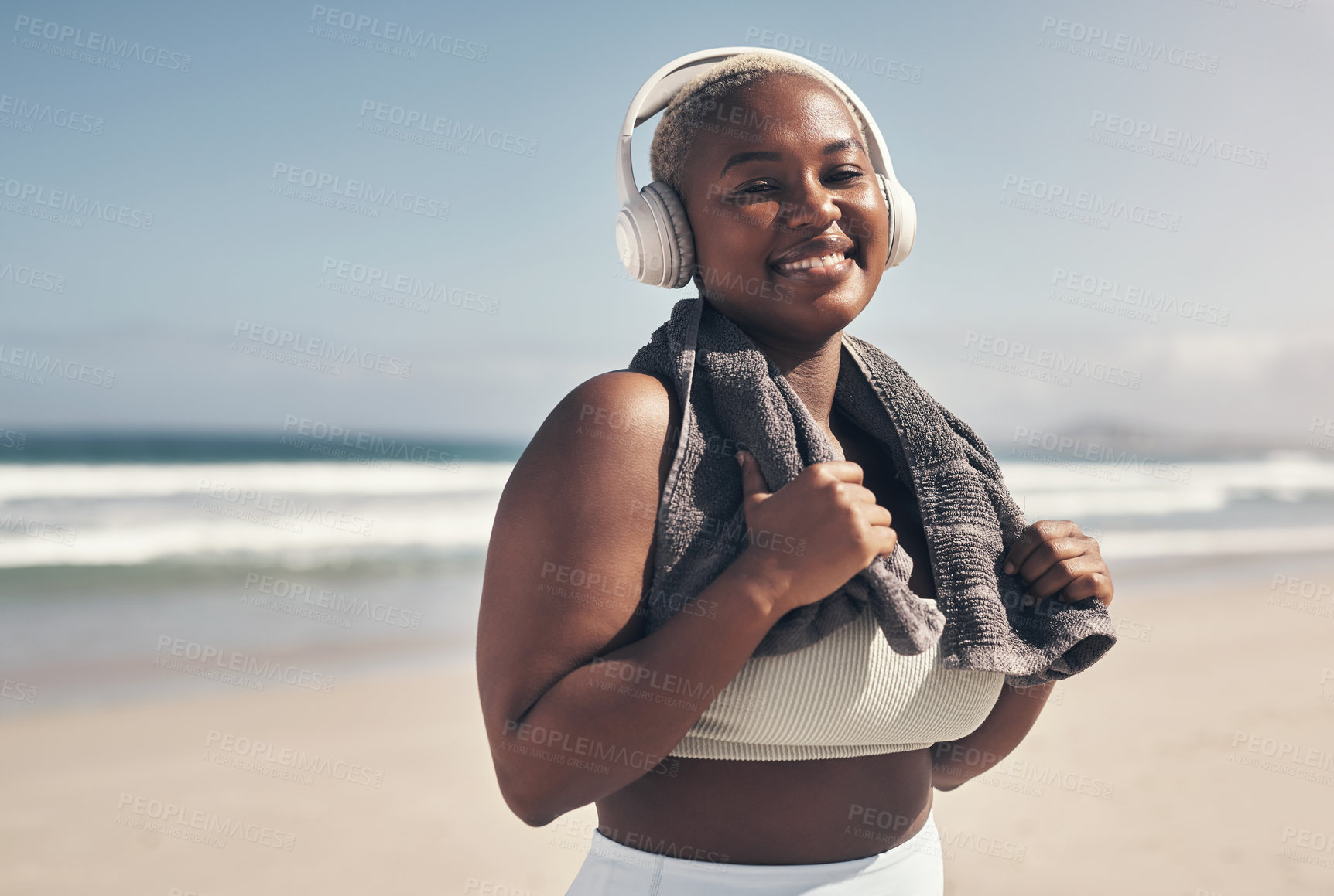 Buy stock photo Shot of a sporty young woman wearing headphones and a towel around her neck on the beach