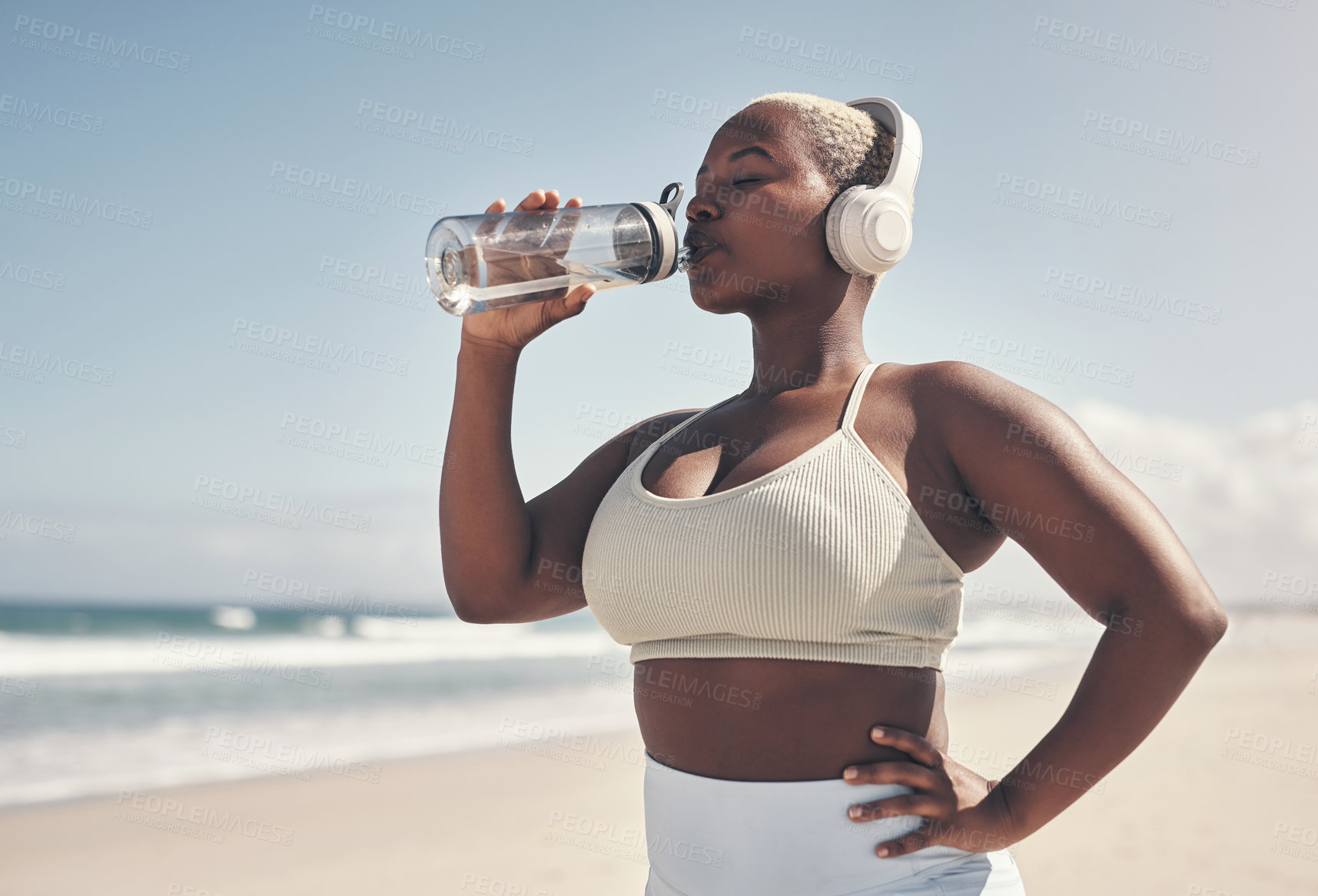 Buy stock photo Shot of a woman wearing headphones and drinking water while out for a workout on the beach