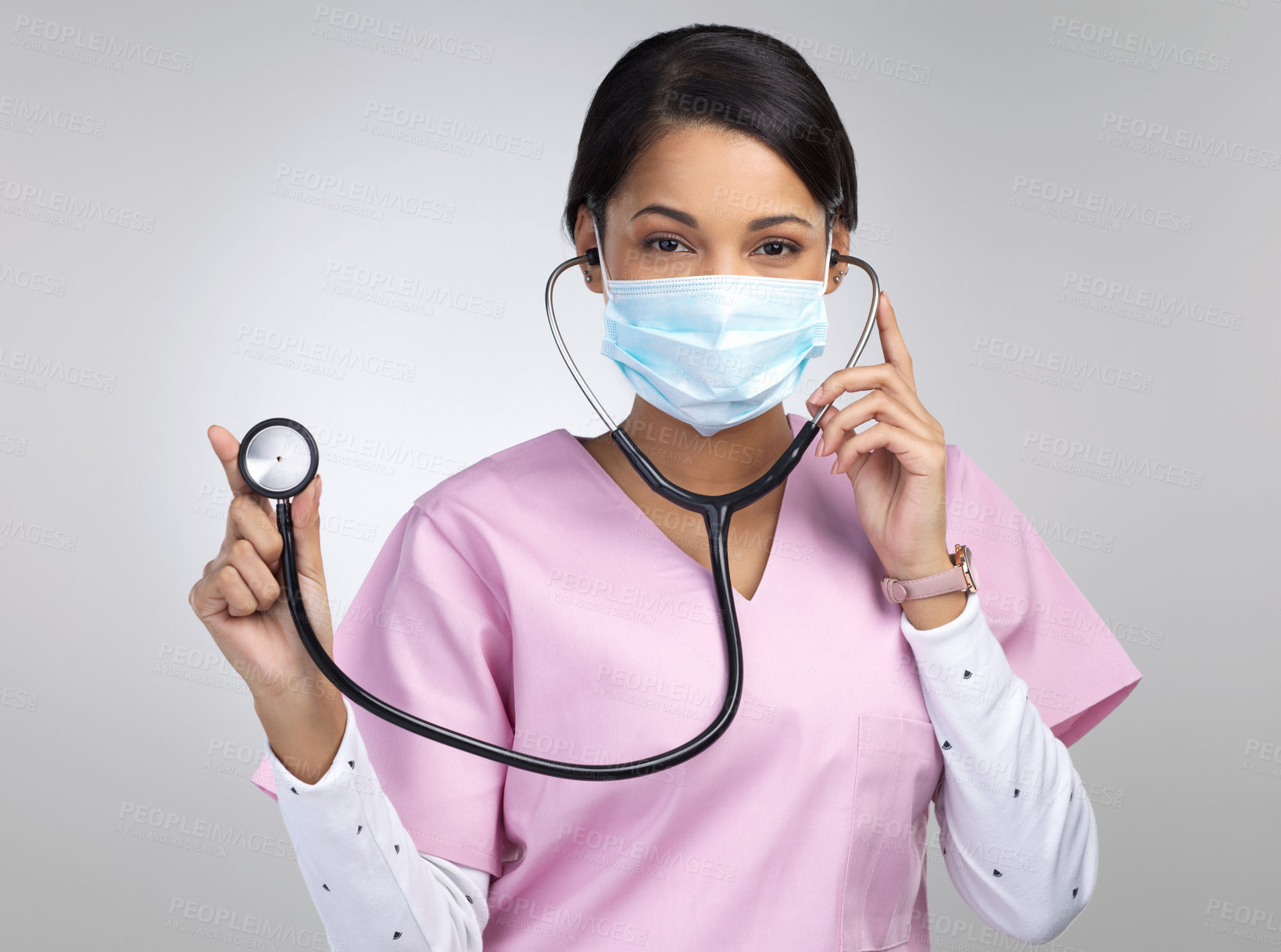 Buy stock photo Cropped portrait of an attractive young female healthcare worker wearing a mask and holding up a stethoscope in studio against a grey background