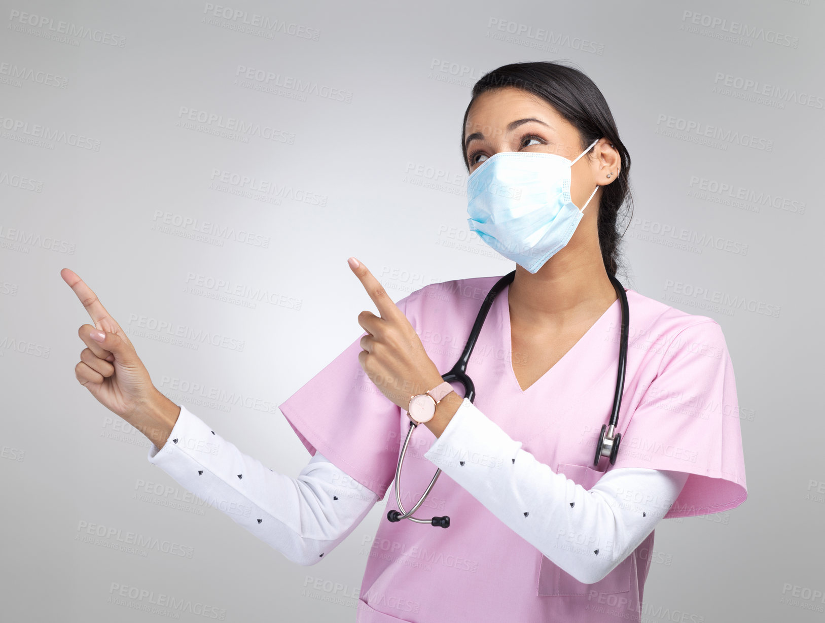 Buy stock photo Cropped shot of an attractive young female healthcare worker wearing a mask and pointing towards copyspace in studio against a grey background