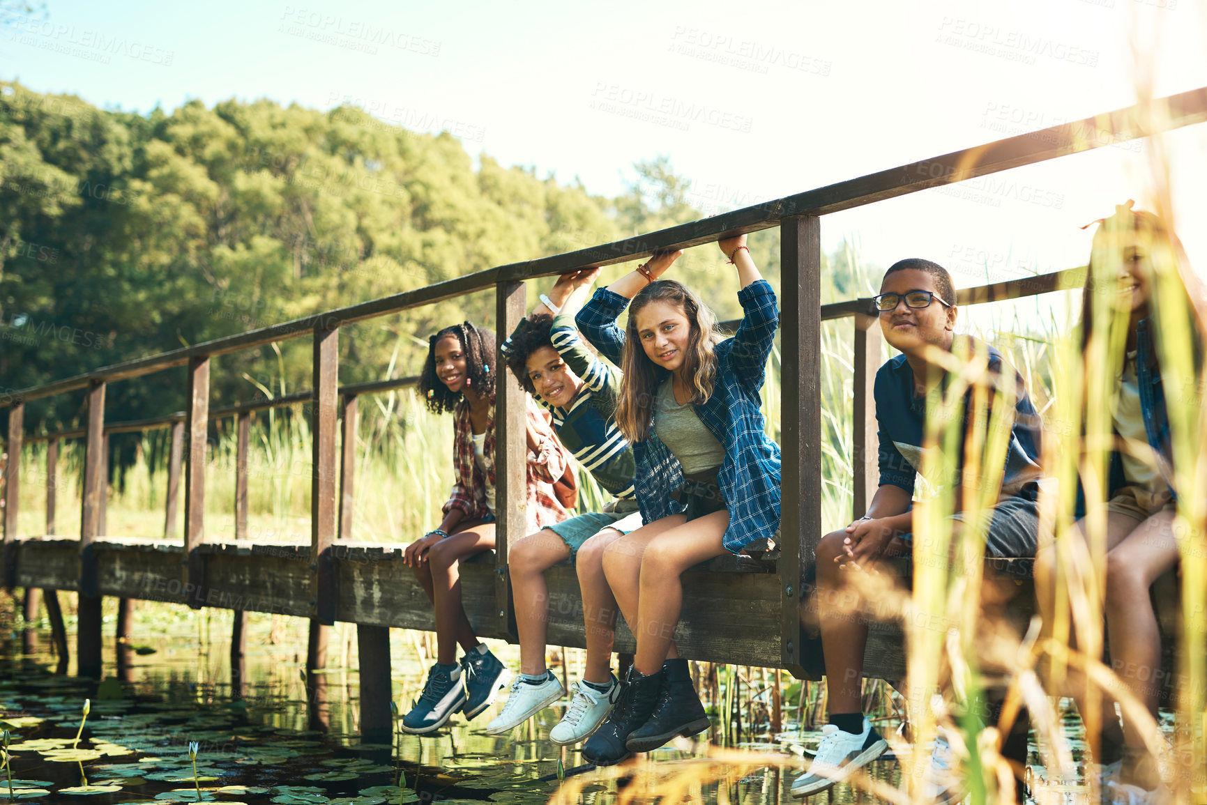 Buy stock photo Shot of a group of teenagers sitting on a bridge in nature at summer camp