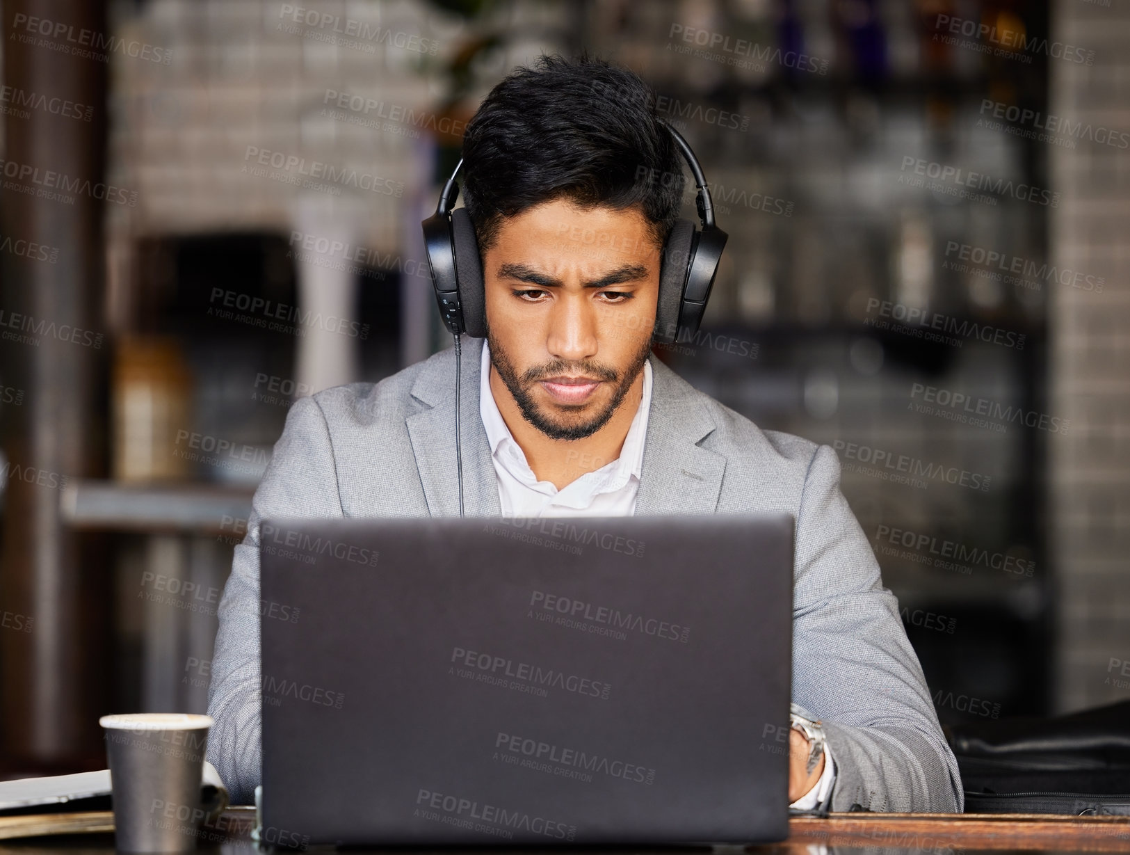 Buy stock photo Shot of a young businessman wearing headphones while using a laptop at a cafe