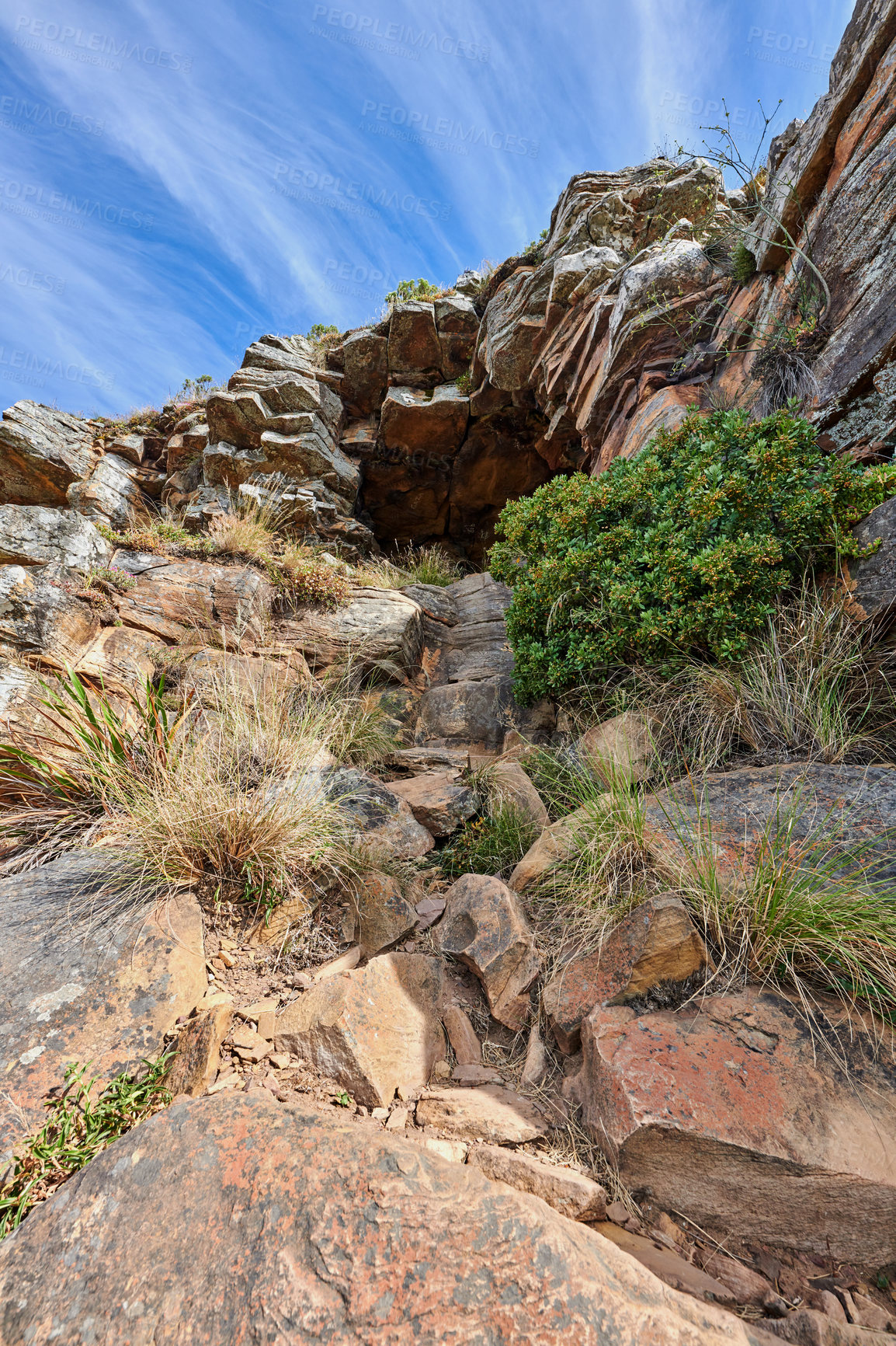 Buy stock photo A rocky mountain with plants and shrubs growing against a cloudy sky background with copy space. Rugged, remote and quiet landscape with rocks and stones on a cliff leading to a mysterious cave