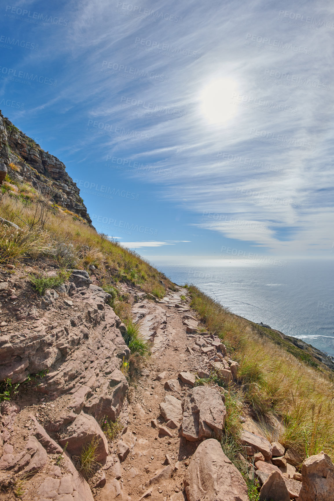 Buy stock photo Secluded mountain trail above a beautiful ocean view and cloudy blue sky. Mountainous walking path surrounded by green grass and rocks. Remote destination to explore and enjoy adventure walks 