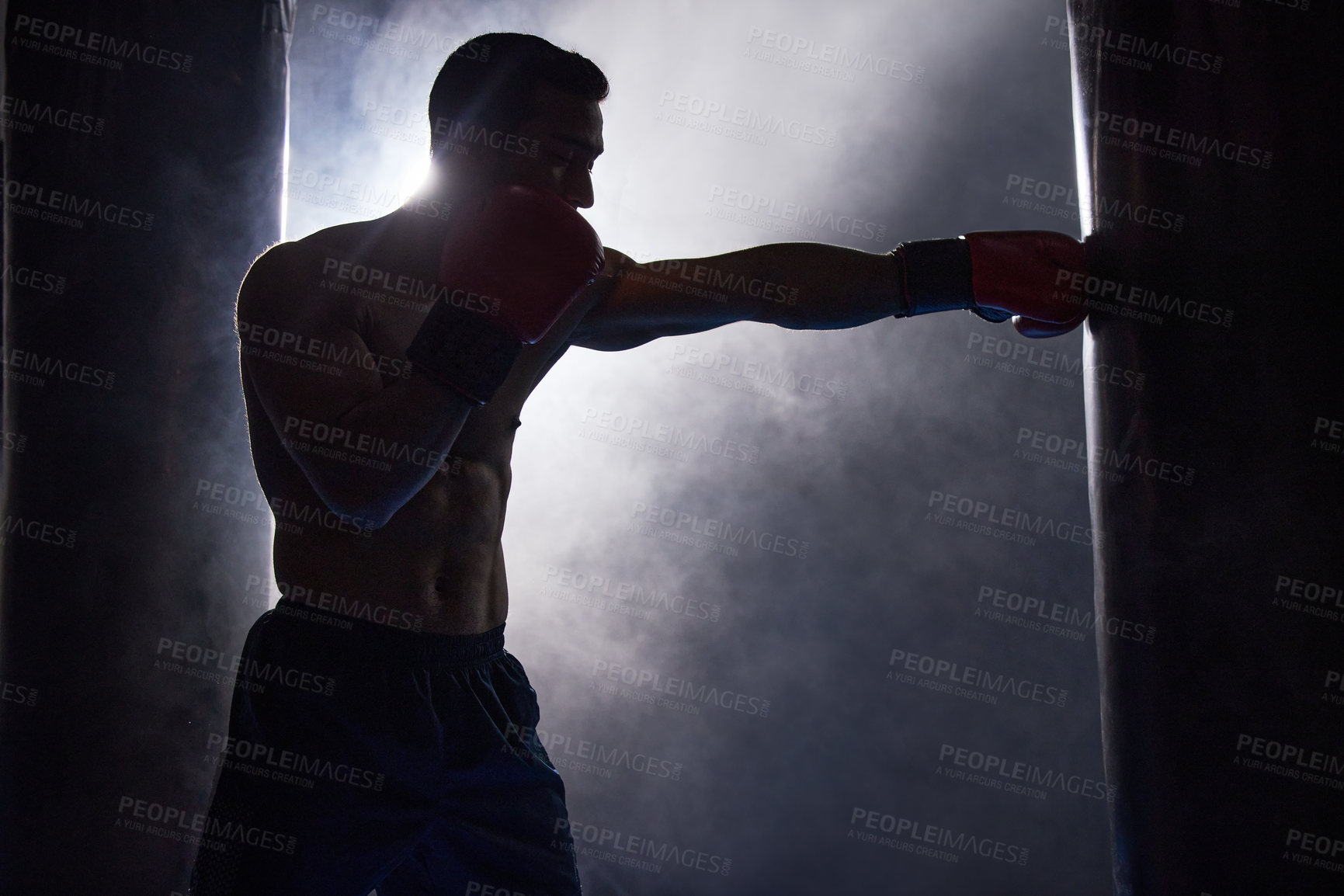 Buy stock photo Cropped shot of a silhouetted young male boxer working out on a punching bag in the gym