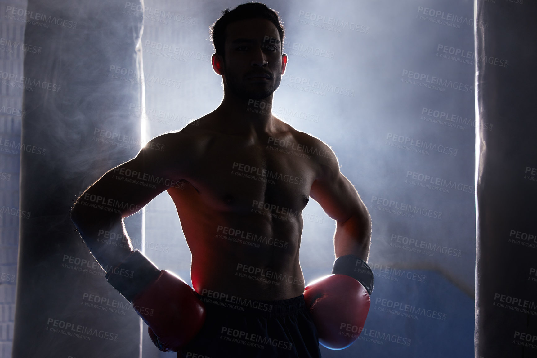 Buy stock photo Cropped portrait of a silhouetted young male boxer standing with his hands on his hips after a workout on the punching