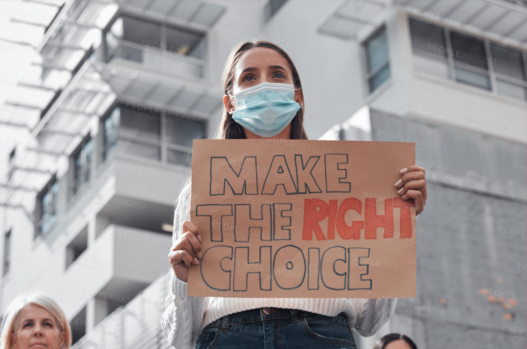Buy stock photo Low angle shot of an attractive young woman holding up a sign protesting against the covid 19 vaccine with other demonstrators in the background
