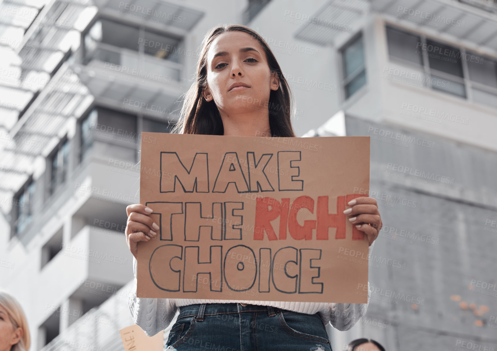 Buy stock photo Cropped portrait of an attractive young woman holding up a sign protesting against the covid 19 vaccine with other demonstrators in the background