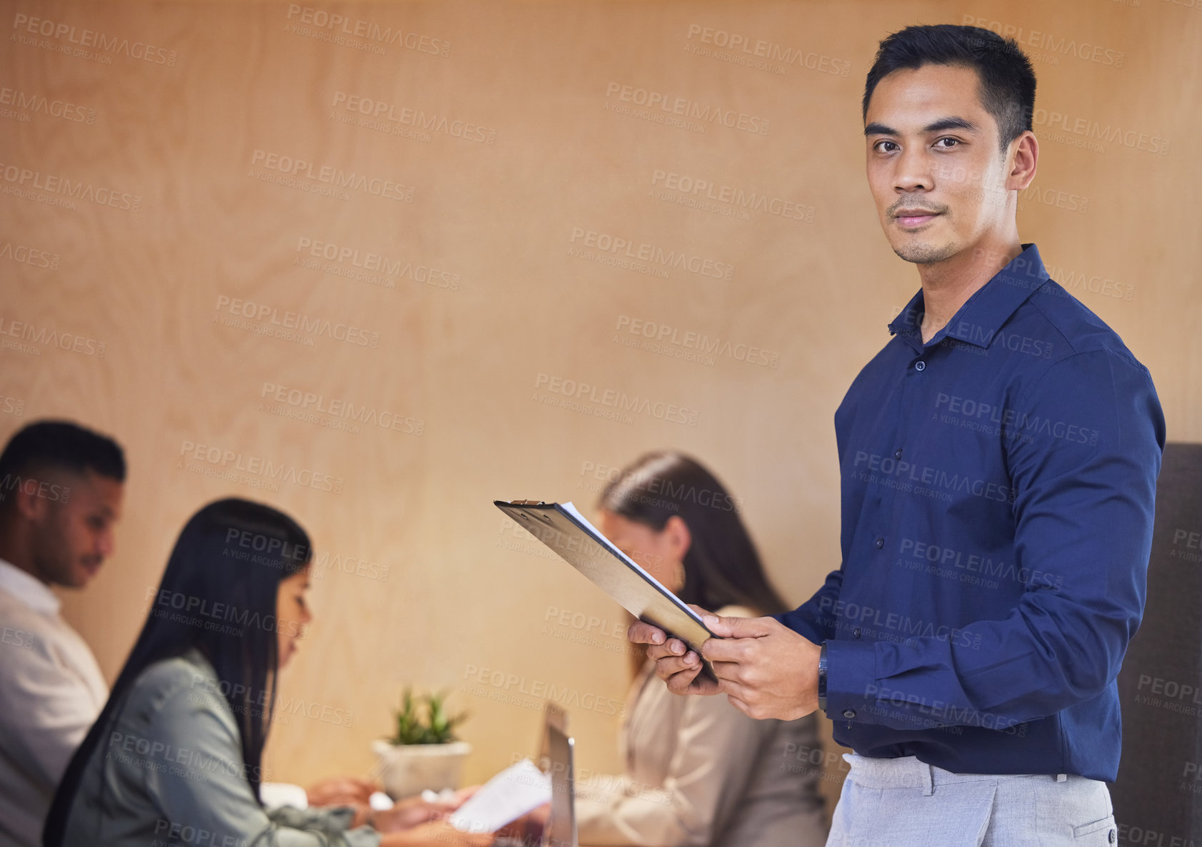 Buy stock photo Cropped portrait of a handsome young businessman standing with a clipboard in the boardroom with her colleagues in the background