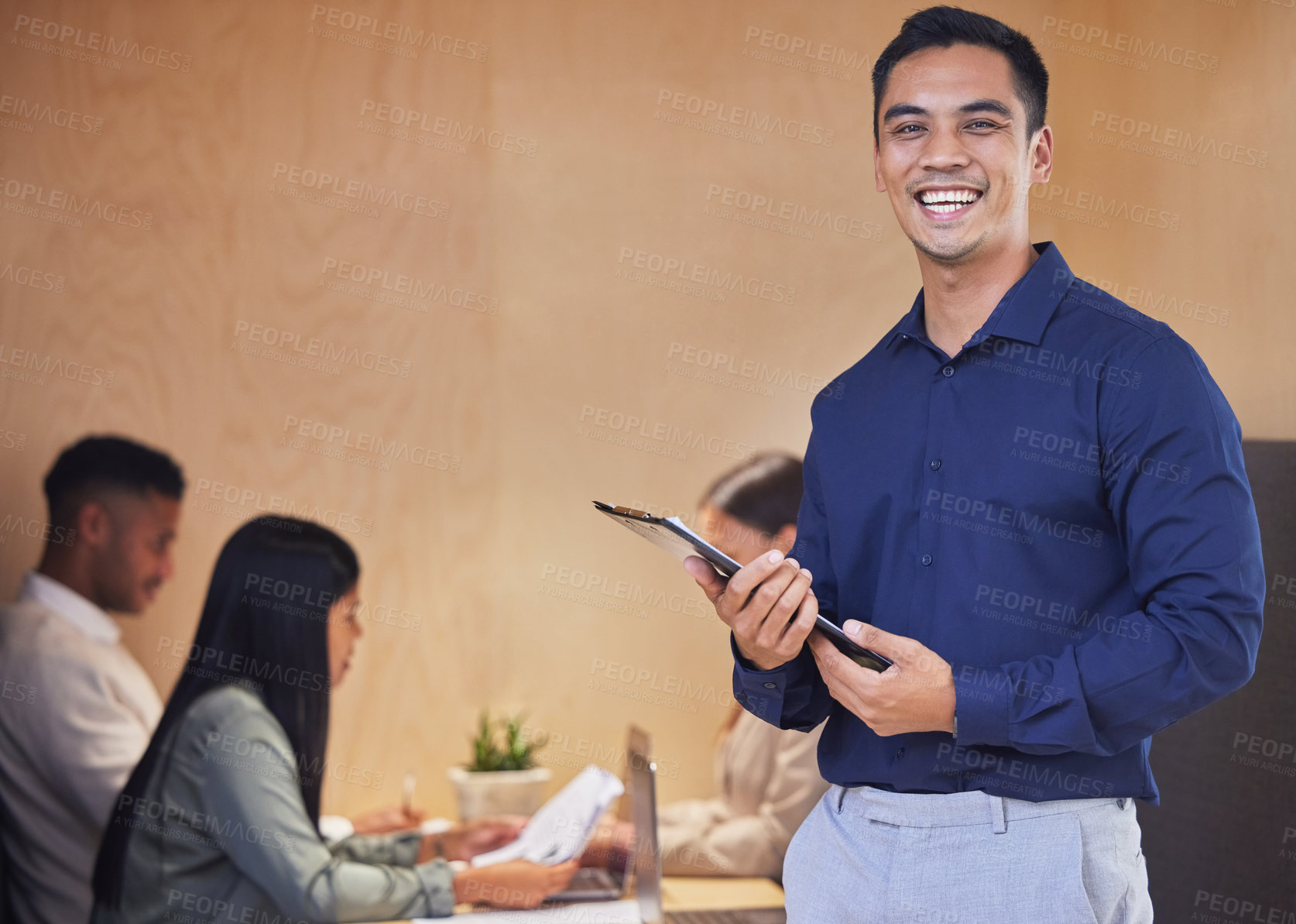 Buy stock photo Cropped portrait of a handsome young businessman standing with a clipboard in the boardroom with her colleagues in the background