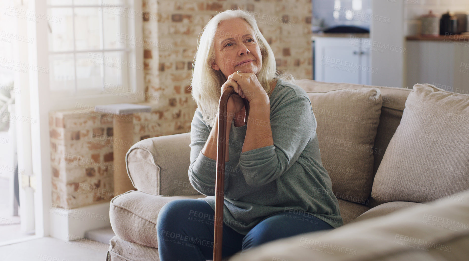 Buy stock photo Shot of a senior woman sitting alone on the sofa at home and looking contemplative while holding her walking stick