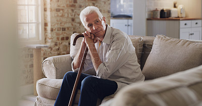 Buy stock photo Shot of a senior man sitting alone on the sofa at home and looking contemplative while holding his walking stick