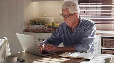 Buy stock photo Shot of a senior man sitting alone in the kitchen and using his laptop to work from home