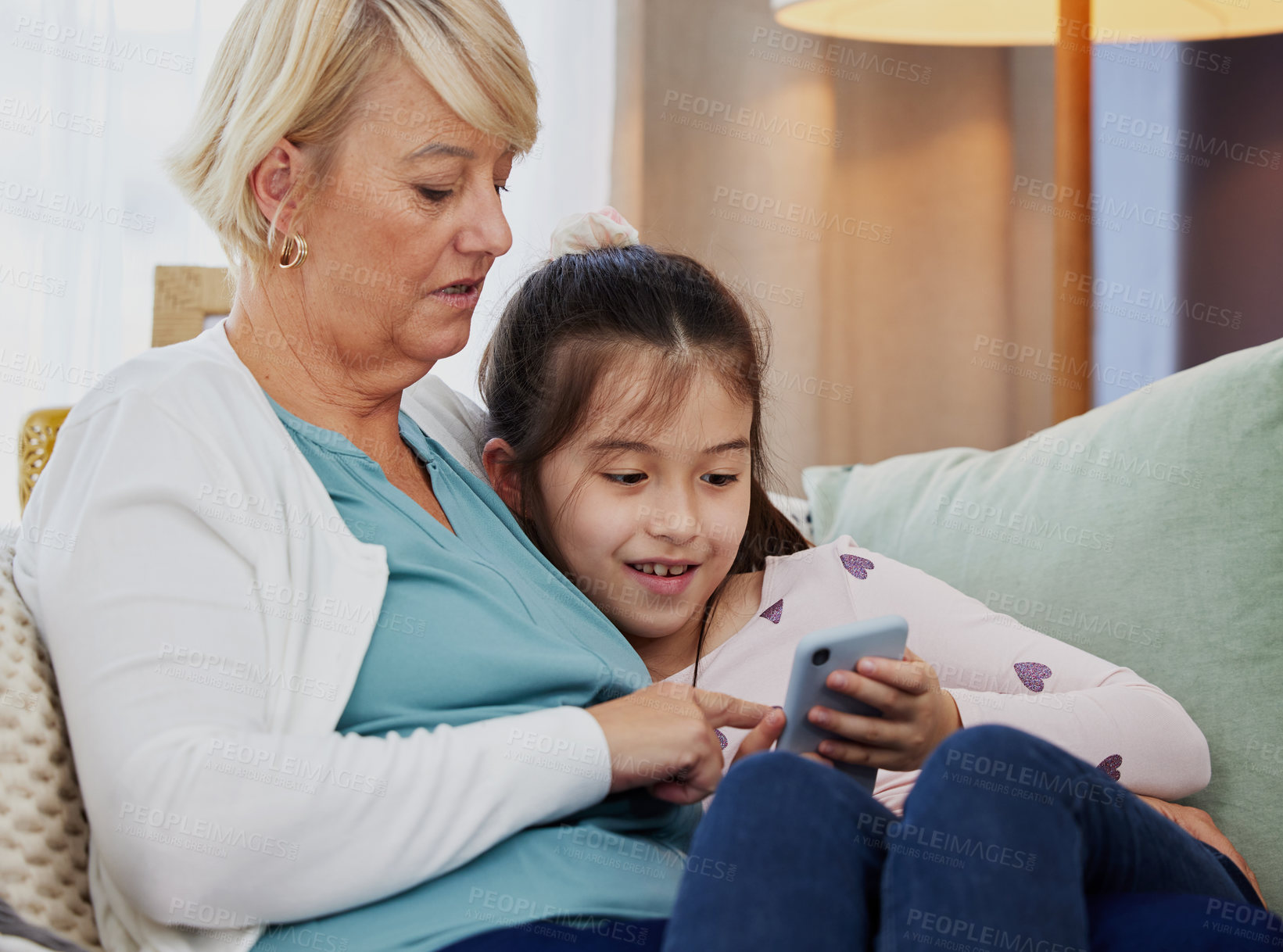 Buy stock photo Shot of a little girl using a cellphone while sitting at home with her grandmother