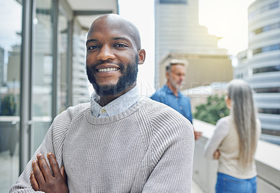 Buy stock photo Cropped portrait of a handsome young businessman standing with his arms folded on the balcony of his office with his colleagues in the background