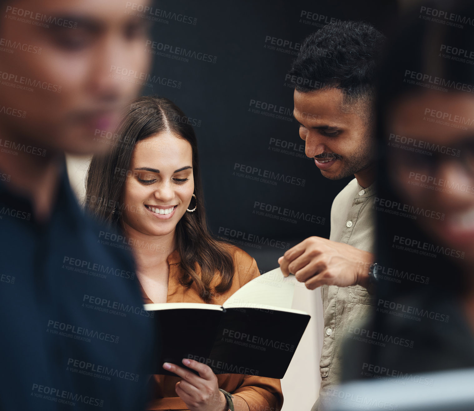 Buy stock photo Cropped shot of two young businesspeople looking over a notebook near the back of their office with colleagues in the foreground