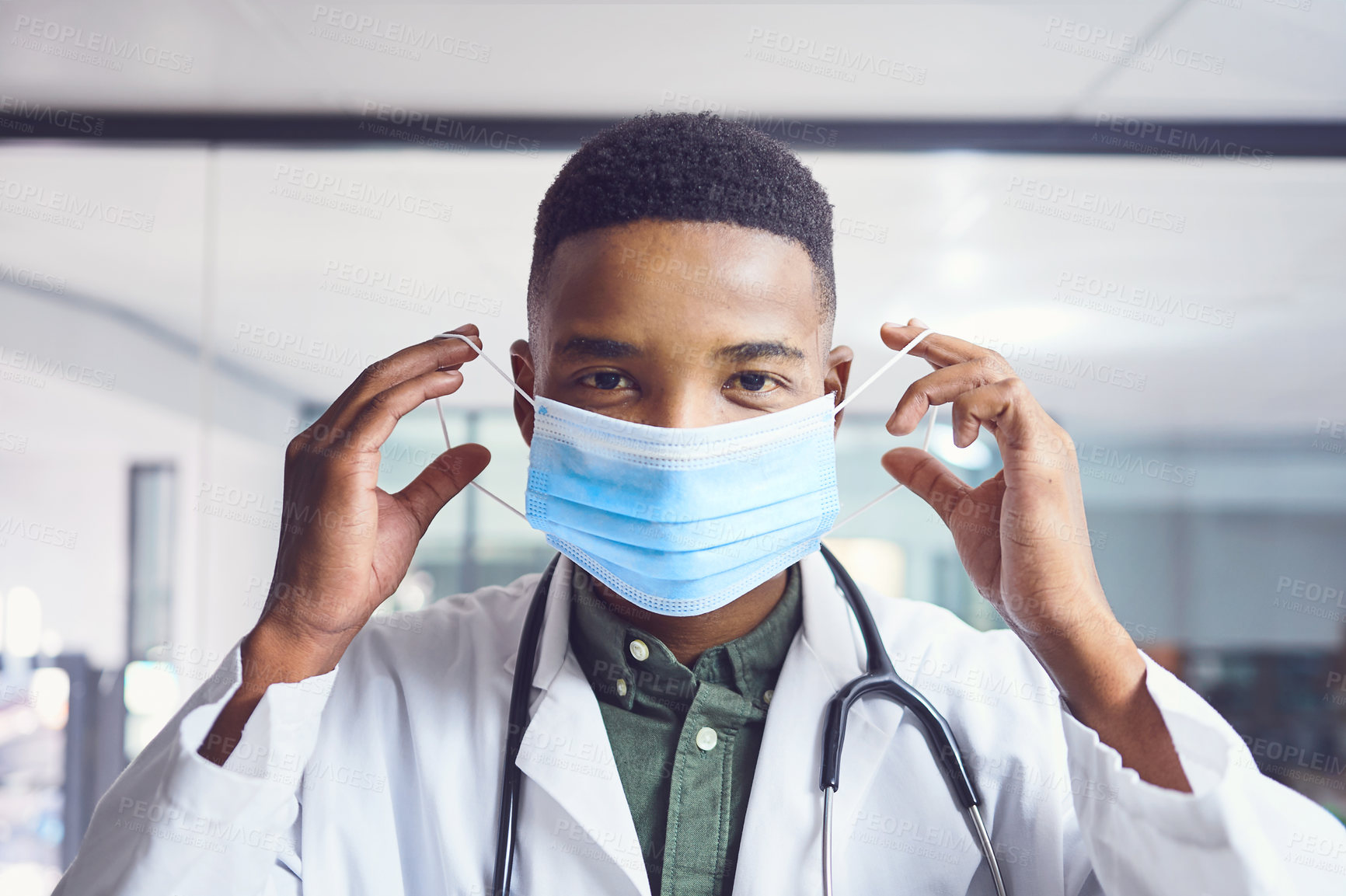 Buy stock photo Cropped portrait of a handsome young male doctor putting on a mask while standing in the hospital