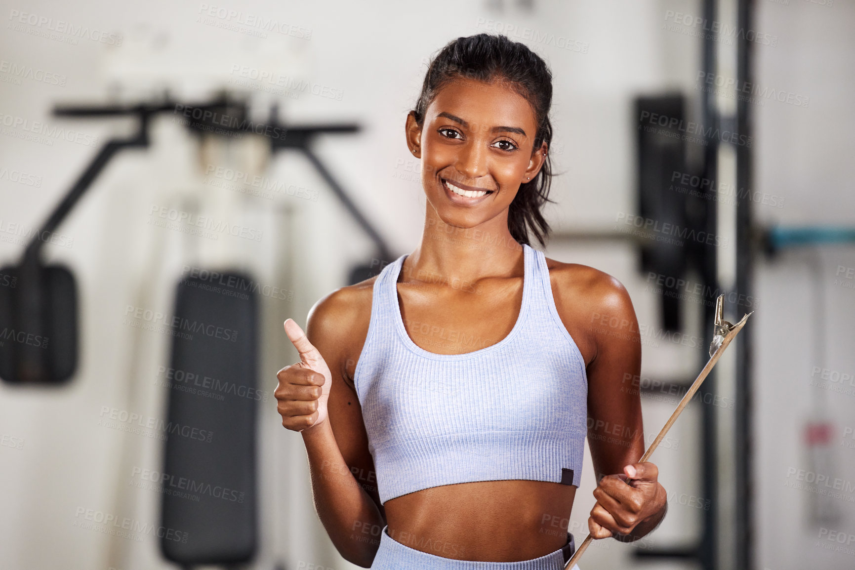 Buy stock photo Shot of a sporty young woman holding a clipboard and showing thumbs up at the gym