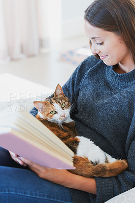 Buy stock photo Shot of a young woman reading a book with a cat on her lap at home
