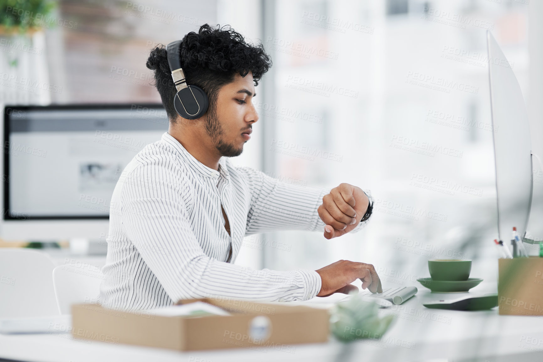 Buy stock photo Shot of a young businessman checking the time on his watch while working on a computer in an office