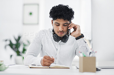 Buy stock photo Shot of a young businessman writing notes while talking on a cellphone in an office