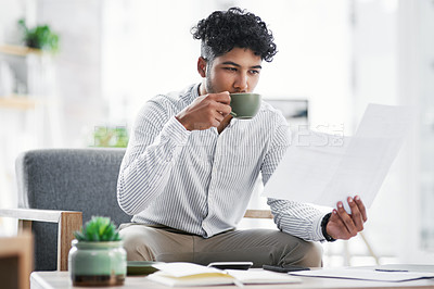 Buy stock photo Shot of a young businessman drinking coffee while going through paperwork in an office