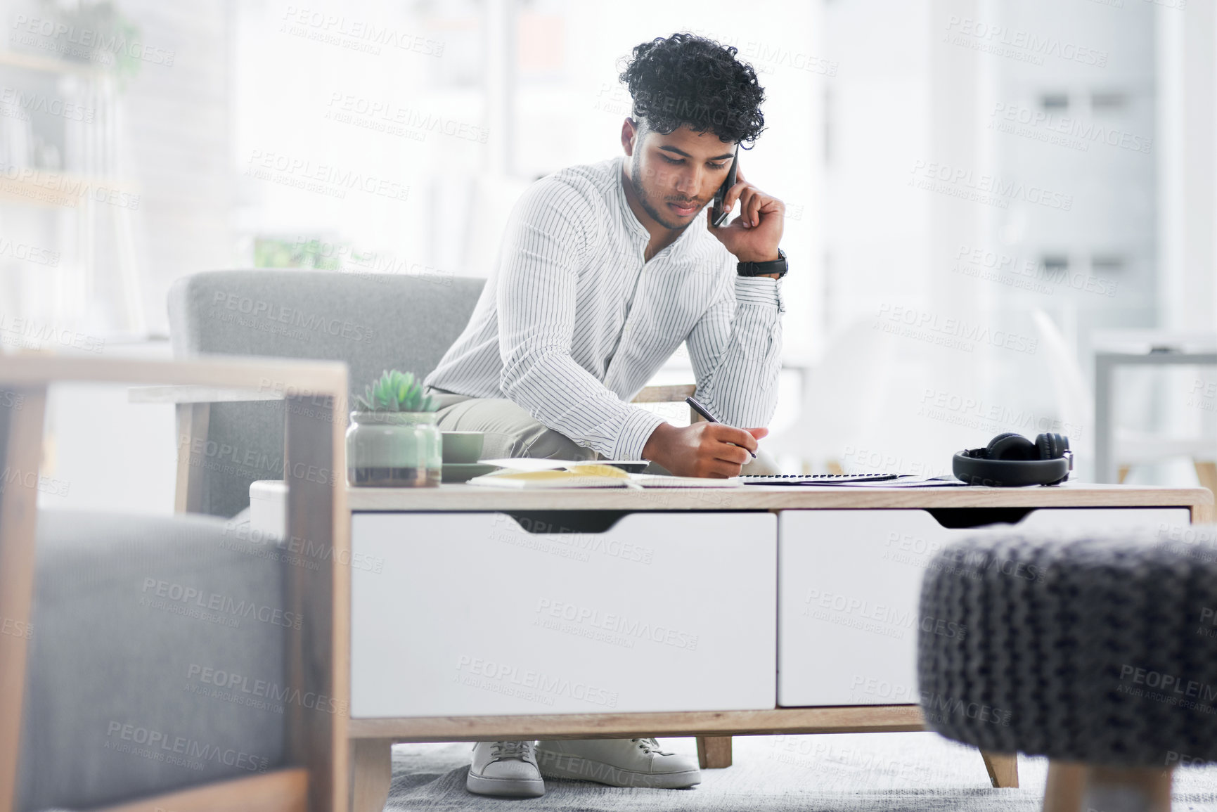 Buy stock photo Shot of a young businessman talking on a cellphone while writing notes in an office