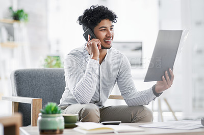 Buy stock photo Shot of a young businessman talking on a cellphone while going through paperwork in an office