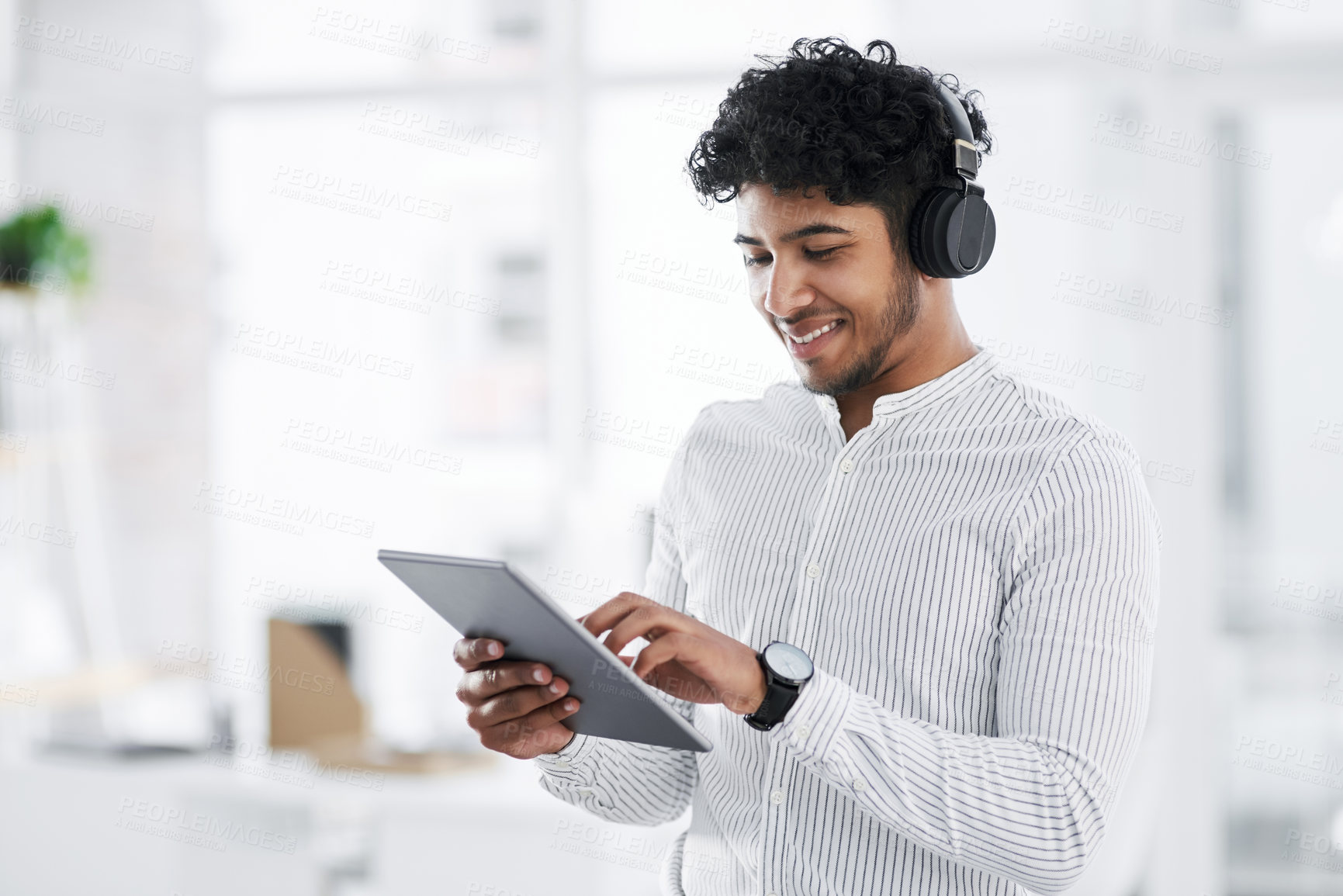 Buy stock photo Shot of a young businessman wearing headphones while using a digital tablet in an office