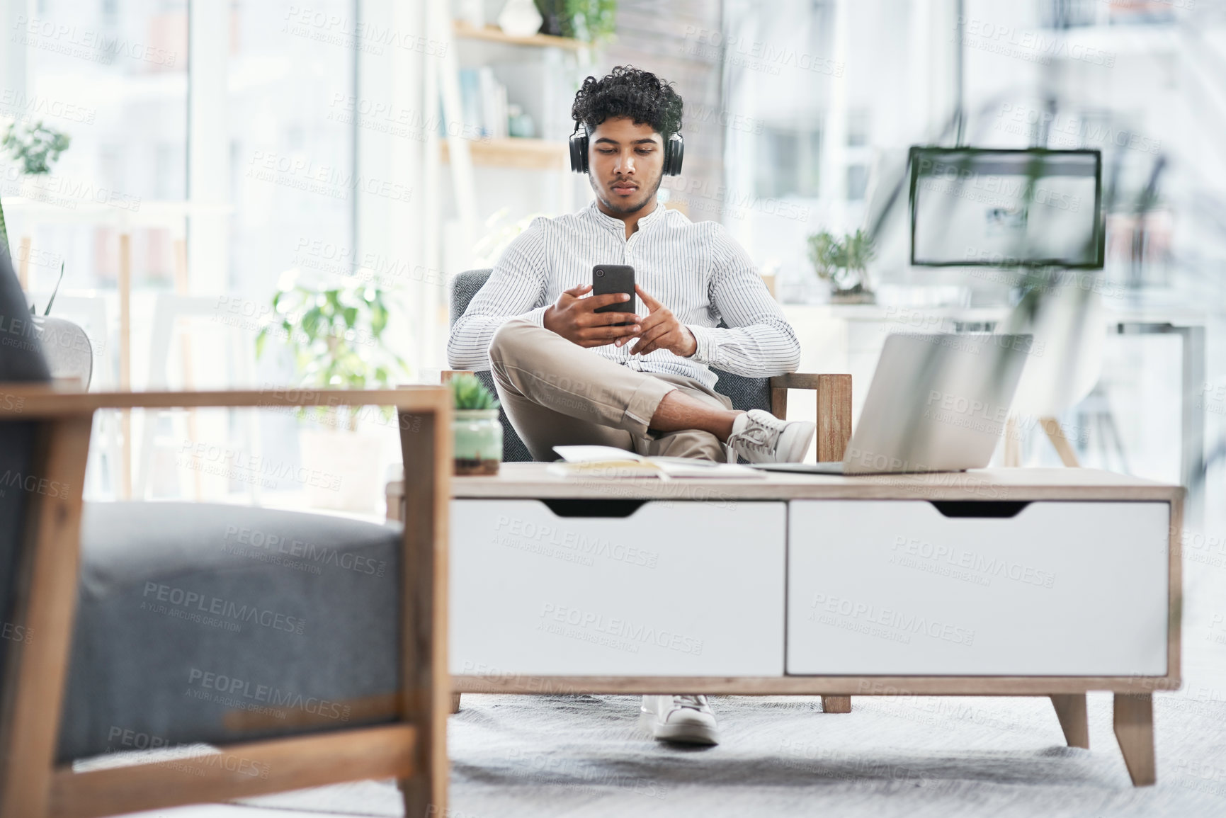 Buy stock photo Shot of a young businessman wearing headphones while using a cellphone in an office