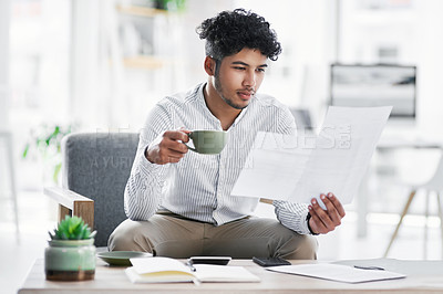 Buy stock photo Shot of a young businessman drinking coffee while going through paperwork in an office