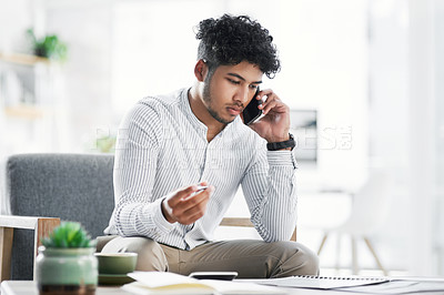 Buy stock photo Shot of a young businessman talking on a cellphone while going through paperwork in an office