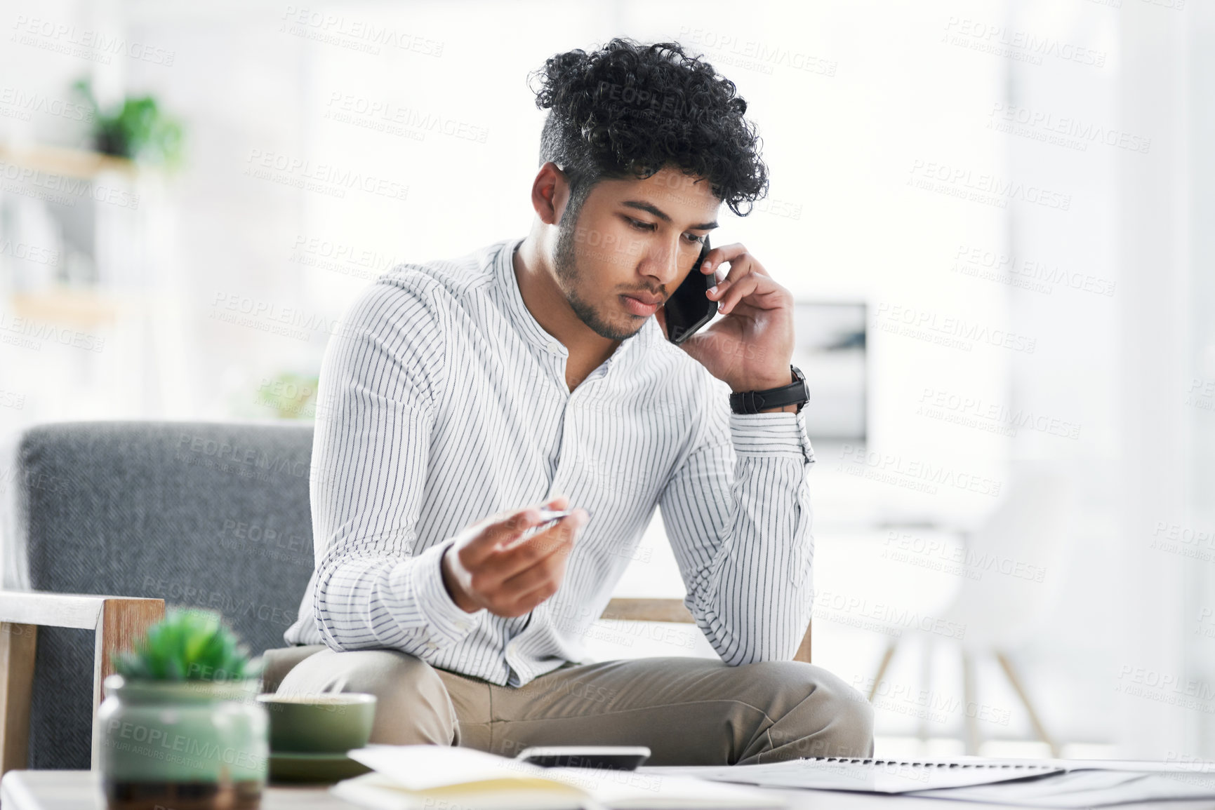 Buy stock photo Shot of a young businessman talking on a cellphone while going through paperwork in an office