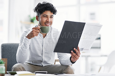 Buy stock photo Shot of a young businessman drinking coffee while going through paperwork in an office