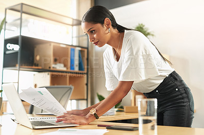 Buy stock photo Shot of a young businesswoman reviewing paperwork at work while using her laptop