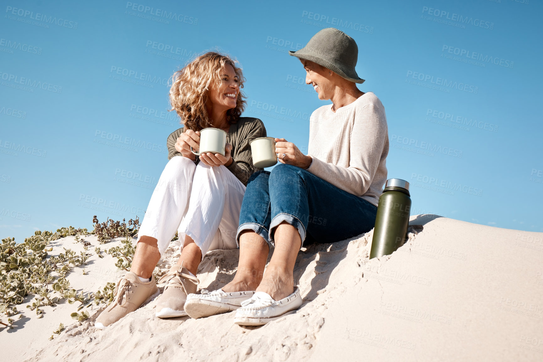 Buy stock photo Full length shot of two attractive mature women enjoying some coffee while sitting on the beach