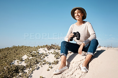 Buy stock photo Full length shot of an attractive mature woman using her binoculars while sitting on the beach