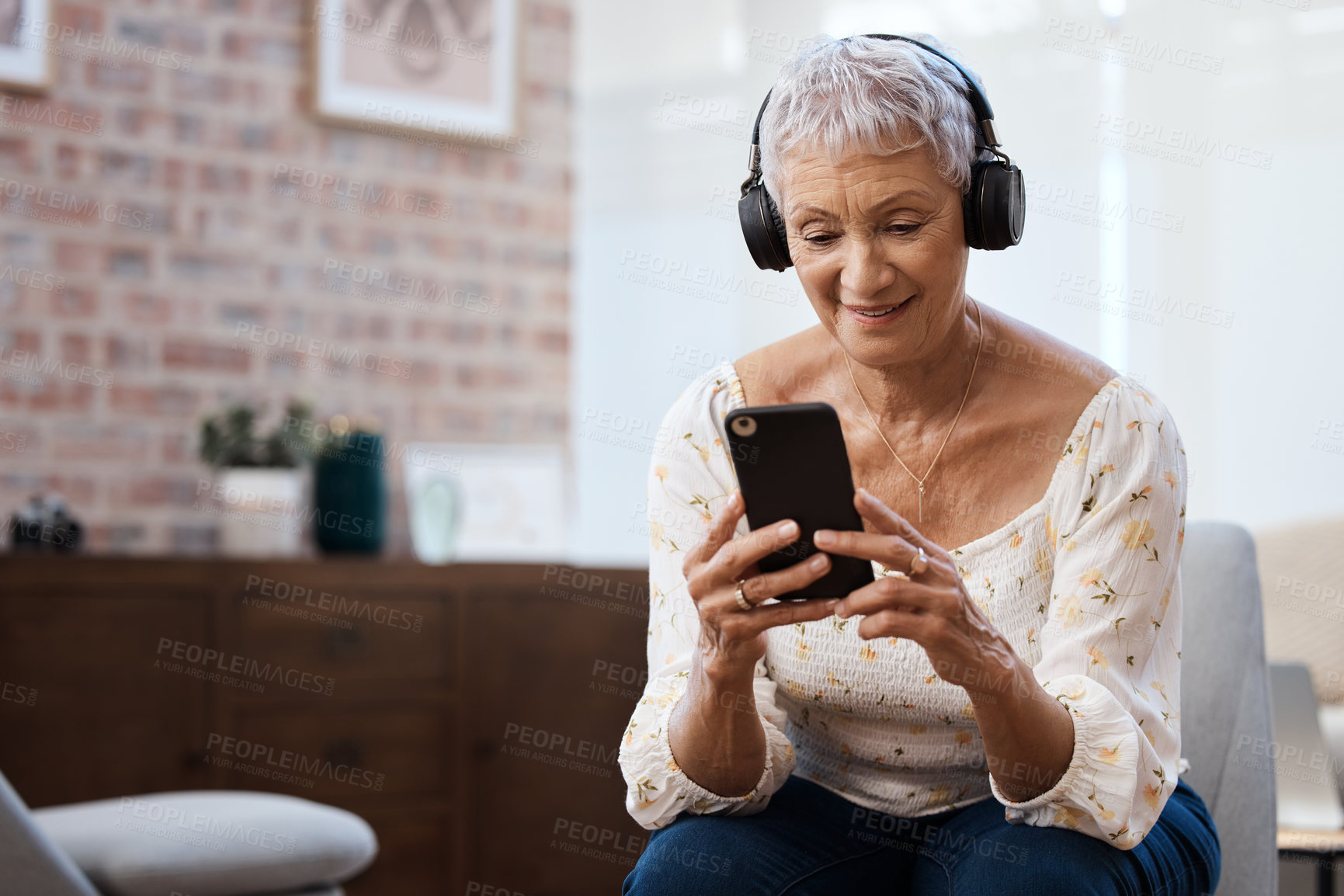 Buy stock photo Shot of a senior woman using a smartphone and headphones on the sofa at home