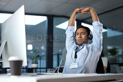 Buy stock photo Shot of a young man stretching while using a headset and computer in a modern office