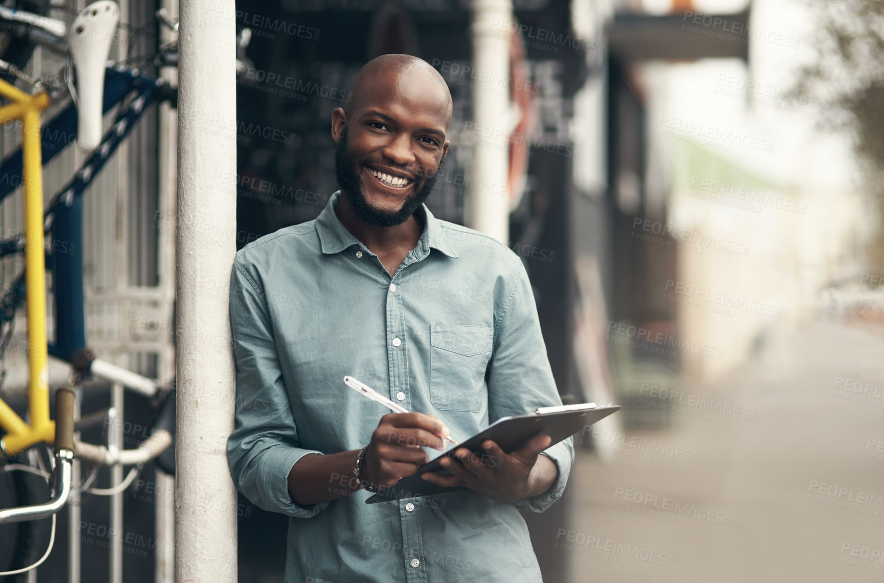 Buy stock photo Shot of a handsome young man standing alone outside his bicycle shop and writing on a clipboard