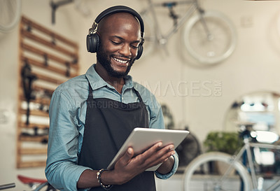 Buy stock photo Shot of a handsome young man standing alone in his bicycle shop and using a digital tablet while wearing headphones