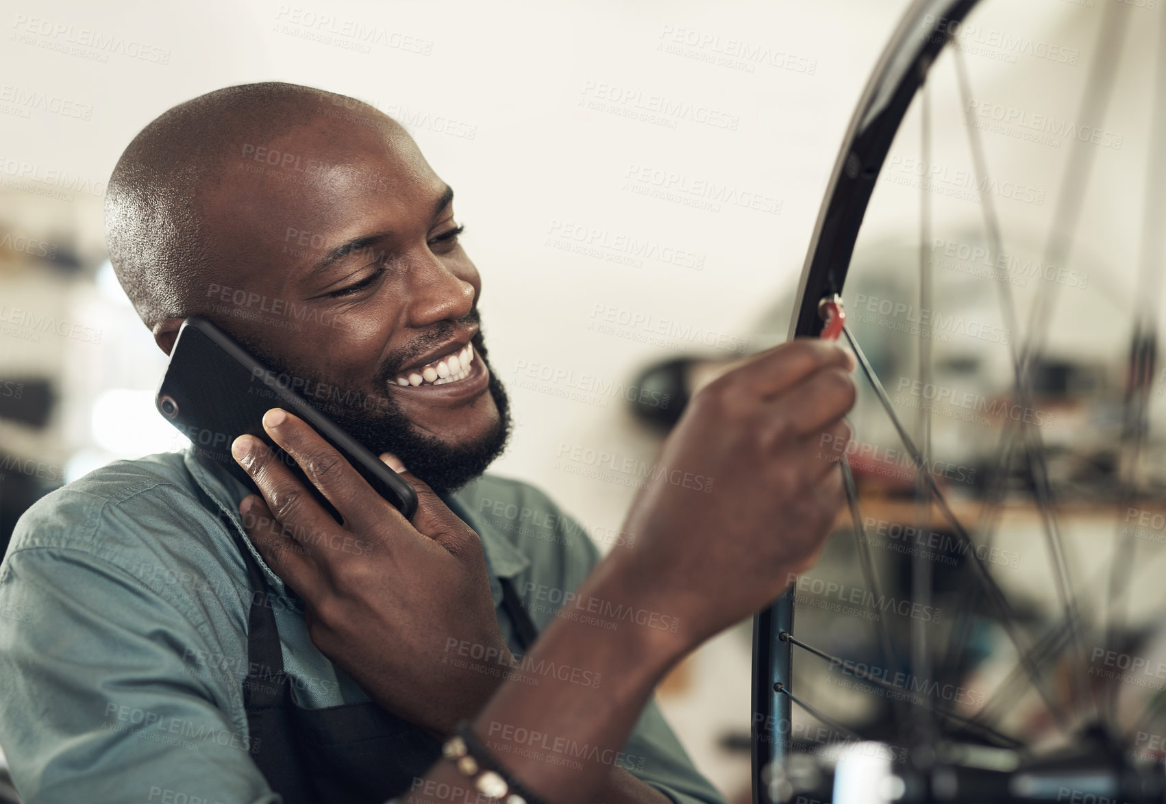 Buy stock photo Shot of a handsome young man standing alone in his shop and repairing a bicycle wheel while using his cellphone