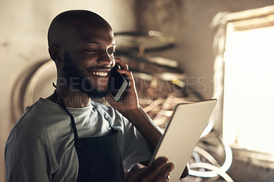 Buy stock photo Shot of a young man talking on a cellphone and browsing on a digital tablet while working at a bicycle repair shop