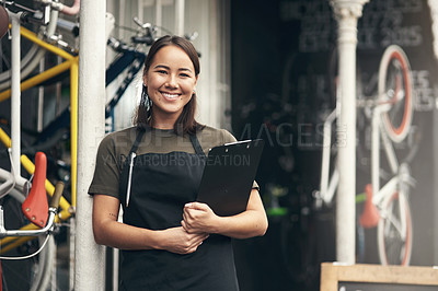 Buy stock photo Shot of an attractive young woman standing alone outside her bicycle shop and holding a clipboard